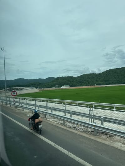 The image depicts a cloudy day in Hoa Xuan, Vietnam, with a person riding a motorcycle on a paved road. The motorcycle is carrying a large bag on its rear rack. In the background, there are lush green rice fields bordered by a metal fence and a highway overpass. Further in the distance, rolling green hills rise under the overcast sky. A speed limit sign indicating "70" is visible on the left side of the road. The scene suggests a rural or semi-rural setting with ongoing infrastructure development, as indicated by the highway construction.