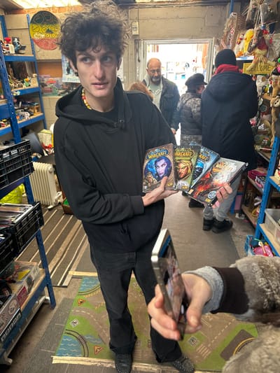 The image depicts the interior of a second-hand goods store or market stall located in Neuilly-Plaisance, France.
A young male, estimated to be in his late teens or early twenties, with dark, curly hair, a pale complexion, and a neutral expression, stands centrally, facing slightly to his left (viewer's right). He is wearing a black hooded sweatshirt, dark jeans, and dark shoes with studs. A multi-colored beaded necklace is visible around his neck. He holds four "World of Warcraft" PC game boxes fanned out in his hands, presenting them towards the camera. The titles visible are "World of Warcraft" (original), "World of Warcraft: The Burning Crusade," "World of Warcraft: Wrath of the Lich King," and "World of Warcraft: Cataclysm."
In the immediate foreground, to the bottom right, a right hand holds a silver-colored smartphone vertically, pointing it towards the young man, indicating a photograph is being taken. The person holding the phone is wearing a grey long-sleeved garment under a dark, possibly faux-fur-lined, outer layer.
The background reveals an open doorway leading to what appears to be an outdoor area or another section of the market. Several individuals are visible in the background: a man with a bald head and grey beard, wearing a light jacket, stands just inside the doorway looking towards the camera; another person, mostly obscured, is behind him; and two more individuals, one in a dark hat and jacket with their back to the camera, are further to the right.
The store environment is densely packed with merchandise. On the left, a blue metal shelving unit contains items like black plastic crates, possibly holding media, and a white radiator is positioned below. On the right, another blue metal shelving unit is heavily laden with various items, including numerous stuffed animals, decorative objects, and a "WELCOME" sign. The floor features a striped beige and brown rug partially covering a colorful children's playmat depicting roads and a river. The ceiling is rough, exposing brickwork or concrete, with visible pipes and wiring, characteristic of an informal retail space.