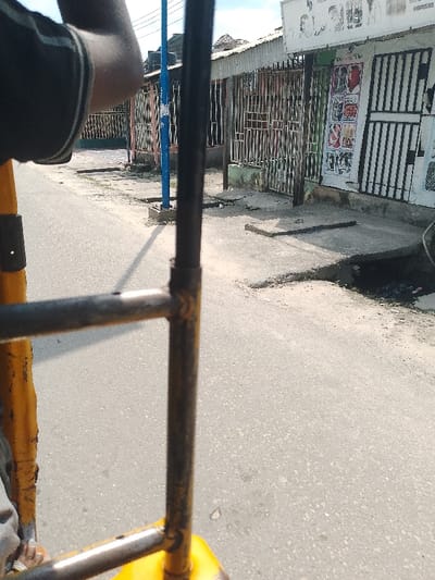 The image presents a street view in Mgbuoshimini, Nigeria, captured from inside a vehicle, likely a tricycle (keke napep), indicated by the yellow and dark metal frame prominent in the foreground. A person's arm, wearing a dark top with a white stripe, is visible in the top-left corner.

The scene depicts a grey, asphalt road that appears well-used and slightly uneven. Bordering the road is a concrete sidewalk with varying levels and a visible open drainage channel on the right, containing dark debris. A flat, square concrete slab rests on the sidewalk further down.

On the right side of the street, a row of single-story buildings, presumably shops, line the road. These structures are characterized by corrugated metal roofs and most have their entrances secured with vertical metal gates or bars. The shop closest to the camera displays a large white signboard featuring black text and images, appearing to advertise hairstyles or beauty products. Below this, a smaller, colorful poster is visible, also depicting what seem to be fashion or beauty-related items.

A tall, slender blue pole stands on the left side of the road. The bright daylight and distinct shadows suggest it is a sunny day, likely during the mid-morning or afternoon. The overall impression is that of a typical street in a commercial or residential area, reflecting local life and commerce. No specific activity beyond being in transit is visible.