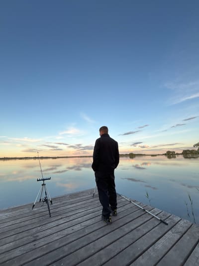 A man stands on a wooden dock, facing the water with fishing equipment nearby. The sky is clear with light clouds reflected on the water's surface. The location appears to be Khryashchevka, Russia.