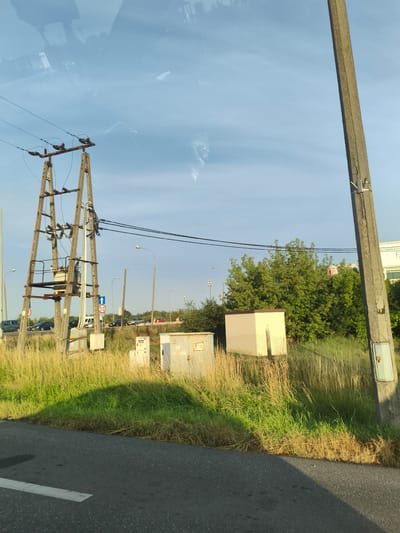 The image depicts an outdoor scene in Cząstków Mazowiecki, Poland, with a focus on infrastructure and a roadside environment. In the foreground, a dark asphalt road with a white lane marker is visible. Tall, dry and green grass lines the edge of the road. Dominating the left side of the image is a tall wooden power pole with multiple crossbeams supporting electrical wires and insulators. A transformer is mounted on this structure. Several smaller electrical boxes or cabinets are situated at the base of the power pole and further into the grassy area.
To the right, another tall concrete pole stands vertically. This pole has a small electrical box attached to its side. In the middle ground, a low, light-colored building or enclosure with a fenced area in front of it is visible, surrounded by more vegetation. Beyond this, a dense cluster of green trees forms a backdrop.
Further into the scene, a road with several cars is present, suggesting a traffic area. Streetlights are positioned along this road. The sky above is a clear blue with some wispy clouds. The overall impression is of a rural or semi-urban area with active electrical infrastructure alongside a road and natural growth. Reflections are visible on the surface from which the photo was taken, possibly a window.