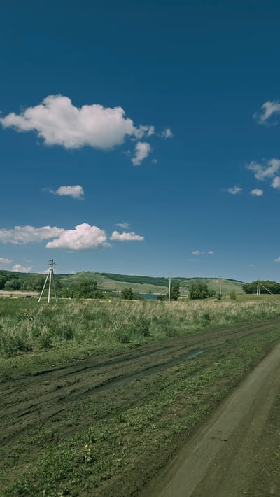 The image depicts a rural landscape under a bright blue sky with scattered white clouds. In the foreground, a muddy dirt road with tire tracks and puddles cuts across a grassy field. Beyond the field, a line of trees and rolling hills stretches across the horizon. Several utility poles with wires are visible in the middle ground. A body of water appears in the distance, nestled among the hills. The overall scene suggests a sunny day in a countryside setting. The provided information indicates the location is the city of Победа, Russia, however, there are no specific identifiable landmarks within the image that would confirm this particular location without further context.