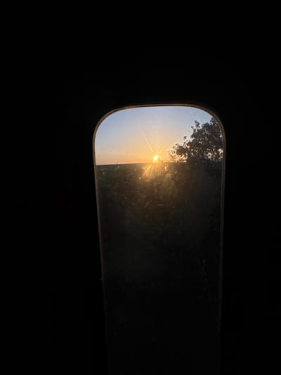 An outdoor scene captures a sunset viewed through a small, arched window. The sun, low on the horizon, casts a warm glow across a field, with a silhouette of trees on the right. The window frame is dark, and the surrounding environment is black, suggesting the image was taken from within a dark interior looking out. The location is Kaspichan, Bulgaria.