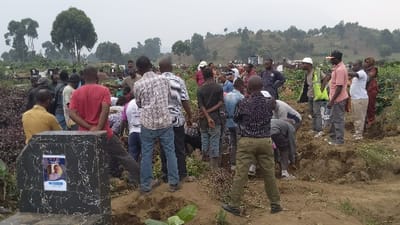 A somber scene unfolds at a cemetery. A crowd of people, mostly men, are gathered around a burial plot. A tombstone with a photo memorial stands nearby. The attendees display a mix of emotions, some looking downcast, others observing the proceedings intently. The background reveals a hillside dotted with trees and other gravesites, creating a landscape of remembrance. The overall atmosphere suggests a funeral or memorial service.