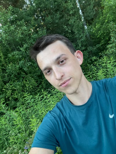 The image is a portrait of a young man taken outdoors in a natural setting, likely a park or garden, as indicated by the abundant green foliage and trees. The man is positioned in the foreground, with his face turned towards the camera. He has short, dark hair and is wearing a teal t-shirt with a subtle white logo, possibly a swoosh, on the upper chest. His skin appears clear, and he has brown eyes and a neutral expression.

The background is dominated by lush green plants and trees, suggesting a daytime setting, possibly late spring or summer. The lighting appears natural, without harsh shadows, implying it could be a slightly overcast day or the image was taken during the golden hour. The overall mood is relaxed and informal. There is no text visible in the image, apart from the logo on the t-shirt. The provided location context is Шевлягино, Russia.