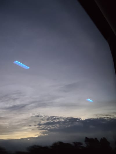 The image depicts a sky with scattered clouds at what appears to be dusk or dawn, with the horizon obscured by dark silhouettes of trees. There are two distinct rectangular, glowing blue lights suspended in the sky. The larger of the two lights is located in the upper left quadrant of the image and appears to be composed of multiple smaller lights or segments. The second light is smaller and is located in the lower right quadrant. The sky has a muted color palette, ranging from light gray to a darker, hazy blue. The overall impression is that of a wide-angle shot looking upwards. No specific location within Malo Buchino, Bulgaria, can be identified from the visual information provided.