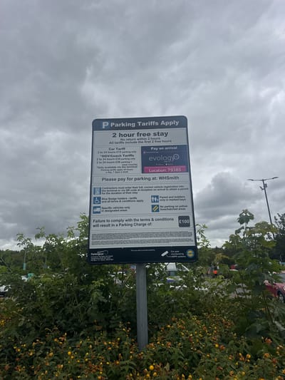 A parking tariff sign is displayed outdoors, mounted on a pole, with lush green bushes and small yellow flowers in the foreground. The sign is blue and white, featuring text about parking rules and charges, including a "2 hour free stay" policy. It mentions "Parking Tariffs Apply," "Please pay for parking at WHSmith," and a potential "failure to comply" charge of £100. The background shows a cloudy sky and a hint of vehicles and a lamppost further back. The sign also includes details for "Parkingeye," a company number, and a PO Box address in Blyth, UK. The location is identified as WHSmith.