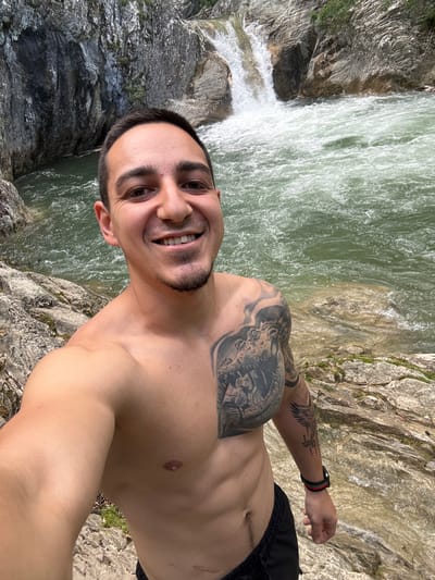A shirtless man smiles directly at the camera, holding it at arm's length. He stands on a rocky outcrop adjacent to a waterfall and a rushing river. The man has dark hair, a trimmed beard, and tattoos covering his chest and upper arm. He wears a wristband on his left wrist and black shorts. The scene appears to be located outdoors, possibly near Medven, Bulgaria, given the implied location. The focus is on the man and the natural backdrop of water and rock.