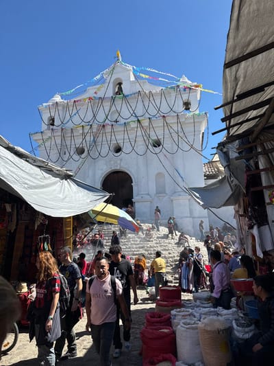 A white colonial church stands tall against a blue sky, adorned with colorful streamers and dark looped decorations. People mill about in front, some on steps leading to the entrance. Market stalls line the edges, shaded by tarps. Bags of goods are visible near the top of the frame. A fair-skinned man in a pink shirt faces the camera in the foreground.