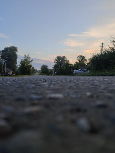 The image shows a low-angle view of a paved road leading through a rural area. On the left side of the road, there are houses and trees. A dark-colored sedan is parked on the side of the road in front of a house, partially obscured by bushes. On the right side of the road, there are more trees and a utility pole with electrical wires. The sky is a pale blue with some wispy clouds tinged with orange from what appears to be sunset or sunrise. The foreground is dominated by the textured surface of the asphalt road, with some small stones visible. The location is identified as Sovkhoz "Kommunar", Russia. There are no people visible in the image.