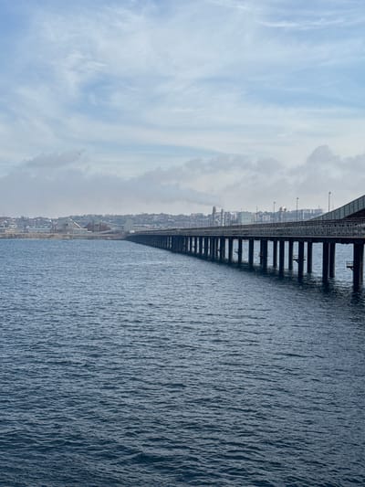 The image depicts a broad, dark blue body of water, likely the sea, with a slightly rippled surface. A large, industrial-looking bridge stretches across the scene, supported by numerous dark, rectangular pylons. The bridge appears to be made of metal and carries a railway line and possibly a road. On the left side of the image, a distant cityscape is visible, characterized by numerous buildings and industrial structures, including what appear to be factories or power plants with tall chimneys. The sky above is a pale blue with wispy, white clouds, suggesting daytime with fair weather. The overall impression is of an industrial or port area. The orientation of the image is sideways.