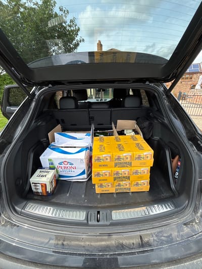 The open trunk of a black SUV is filled with cases of beer, including Wingman Session IPA, Peroni Nastro Azzurro, and Shore Leave. The car is parked in front of a house with a brick exterior, and the scene is captured from a slightly elevated perspective. The coordinates indicate the location is in the city of Newton upon Derwent, United Kingdom. The SUV's rear seats are down to maximize space for the beer cases. There are no people present in the scene.