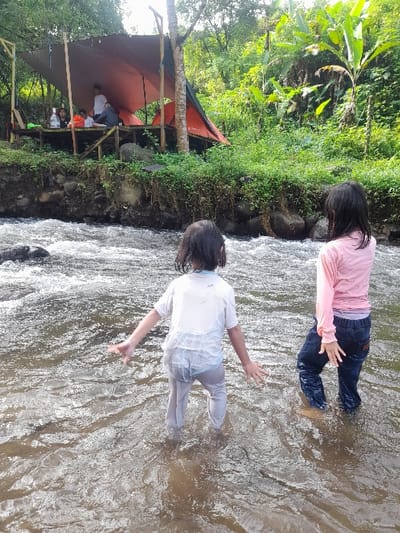 Two young people are wading in a shallow, flowing river. One wears a pink shirt and dark pants, while the other is in a white top and light pants. Both have their arms outstretched. On the riverbank, a makeshift shelter stands with people sitting inside. Greenery and trees surround the area, suggesting a natural setting. The water appears clear, and the sunlight creates bright reflections on the surface.