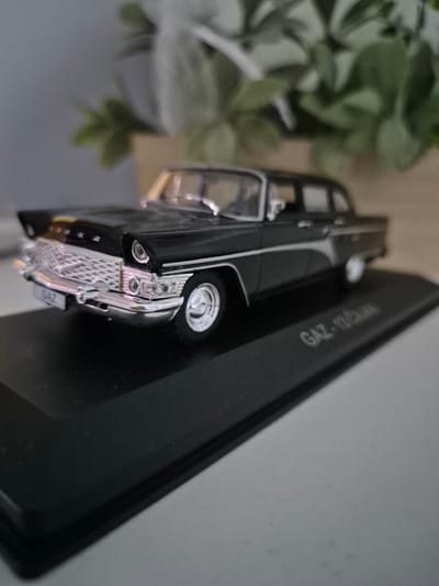 A black die-cast model of a GAZ-13 Chaika sedan is displayed on a black base. The model car is detailed with a chrome grille, headlights, and a license plate that reads "GAZ". The base has the inscription "GAZ-13 Chaika" on its surface. In the blurred background, there is a hint of greenery from a potted plant. The scene is lit with soft lighting, creating reflections on the car's surface. This scene does not depict the city of Iskrets, Bulgaria.