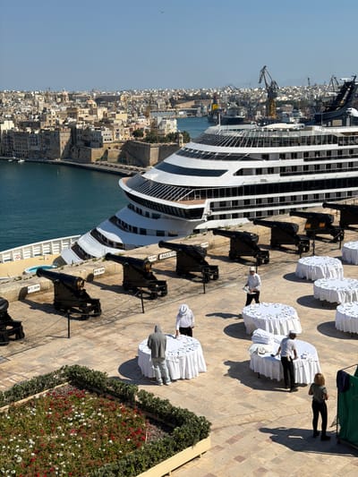 This media file shows an overhead view of a large cruise ship docked in a harbor in Valletta, Malta. The ship's hull is white with black-tinted windows. A cityscape with dense, light-colored buildings is visible behind the ship, suggesting an urban port.
On the paved quayside next to the ship, several round tables are set up, covered with white tablecloths. People dressed in white shirts and dark pants appear to be setting up or attending to the tables, possibly for an event. In the foreground, there are several black cannons mounted on wheeled carriages, arranged in a line along the waterfront. Some of the cannons have small white signs with text. One sign clearly reads "N.7. GUN".
The scene is bathed in bright sunlight, casting long shadows, indicating it is likely daytime, possibly late morning or early afternoon. The sky is clear and blue, suggesting fair weather. The overall atmosphere appears orderly and functional, with a hint of historical ambiance due to the presence of the cannons and the traditional architecture of the city.
