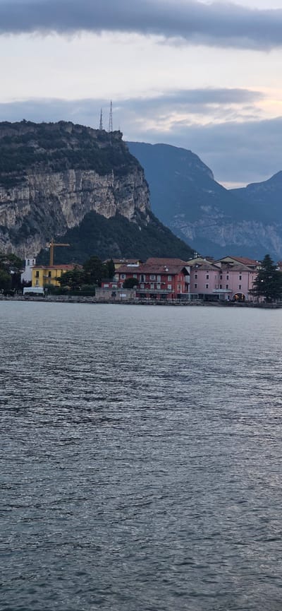 The image captures a serene waterscape in Torbole, Italy, likely during dusk or early morning given the soft, diffused lighting and the cloudy sky. In the foreground, the dark, rippling surface of a body of water, possibly Lake Garda, dominates the lower half of the frame. The water exhibits a textured appearance with consistent, subtle waves reflecting the subdued light.
Along the shoreline in the middle ground, a cluster of buildings forms a picturesque townscape. To the left, a prominent bright yellow building stands out, featuring several windows and a white steeple-like structure attached. A yellow construction crane is visible behind this building, suggesting ongoing development or renovation. Adjacent to it are other structures with red tiled roofs, typical of Italian architecture. One building is painted in a deep red hue, while another adjacent structure displays a light pink facade. These buildings appear to be hotels or residential properties, with visible windows and balconies. A low stone wall or embankment lines the edge of the water in front of these buildings.
In the background, a dramatic, rugged mountain rises steeply, its rocky face showing distinct layers and textures. The mountain is partially covered in dark green foliage, particularly on its lower slopes. At the very peak of this mountain, two tall, slender telecommunication towers are visible against the sky, with one appearing slightly taller than the other. Further in the distance, additional hazy mountain ranges recede into the background, their forms softened by the atmospheric conditions. The sky is overcast with layers of grey and white clouds, with a hint of lighter sky visible towards the right horizon, suggesting the approaching or receding light of dawn or dusk. No people are discernible in the image. The overall impression is one of a tranquil, scenic, and slightly dramatic natural and urban landscape.
