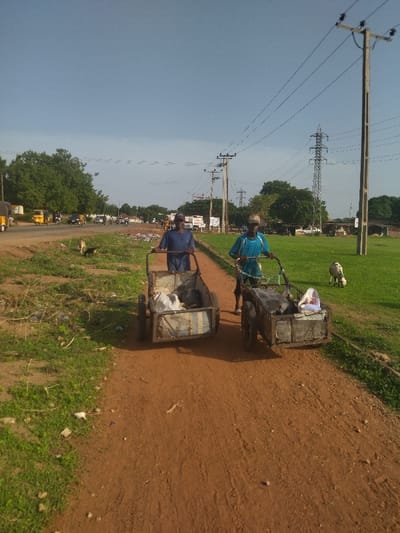 The image shows two men pushing handcarts filled with refuse along a dirt road in a rural area near the city of Kanshio, Nigeria. The location is near the coordinates 7.682, 8.537. Both men are dressed in casual attire, one in a blue shirt and the other in a blue and white striped shirt. There are power lines running overhead and a few trees in the background. The dirt road is dusty, and the grass is green and dry. There is a goat on the grass to the right of the road. The scene appears to be captured on a sunny afternoon. The handcarts are both metal, have two wheels, and are filled with refuse. There are several bags and objects in the handcarts. The men appear to be working, although it is not clear whether they are collecting the refuse or transporting it. The image is a snapshot of everyday life in a rural part of Nigeria.
