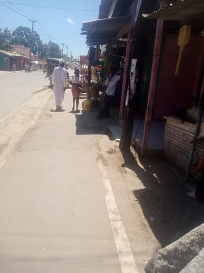The image depicts a daytime street scene in Mtopanga ward, Kenya, under a clear blue sky with high sun casting distinct shadows. The foreground shows a paved road on the left, bordered by a concrete curb and an unpaved dirt sidewalk. On the right, a row of informal roadside shops or stalls is visible, featuring corrugated metal awnings and various merchandise displayed, including bundles of brooms and yellow buckets on the ground.
Three individuals are prominent:
1. A male, dressed in a long white garment and white cap, walks away from the camera on the paved section of the sidewalk.
2. A female, wearing a white top and a reddish-brown knee-length skirt, walks slightly behind and to the left of the male, also moving away from the camera.
3. A male, wearing a light-colored shirt and dark trousers, leans against a dark support pillar of a shop on the right, facing towards the street.
Further down the street, utility poles with overhead wires are visible, along with additional buildings, trees, and distant figures. An auto-rickshaw (tuk-tuk) is also discernible in the mid-ground. A large yellow, paddle-shaped object hangs from one of the shop structures on the far right. The environment is dusty, with sparse vegetation along the dirt path.