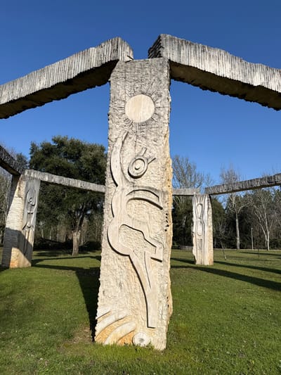 The image displays a large-scale concrete sculptural installation located in Mealhada, Portugal. A prominent, vertical concrete pillar in the foreground exhibits a rough, textured surface with deeply carved relief patterns. These carvings include a circular sun-like motif at the top, a stylized human-like figure in a dynamic pose in the middle, and abstract organic shapes near the base. This central pillar is structurally integrated into a larger framework via two horizontal, ribbed concrete beams extending outwards, connecting to other similar concrete pillars. Two additional, distinctly carved concrete pillars are visible further into the scene, forming part of the broader structure. The entire installation stands on a field of short, green grass, which is marked by strong, elongated shadows cast by the concrete elements, indicating direct sunlight from the upper left. The background features a mix of distant trees, some with bare branches, suggesting deciduous species, and others with dense green foliage, characteristic of evergreens. The sky above is clear and uniformly blue. No individuals, animals, or other moving objects are present in the frame.