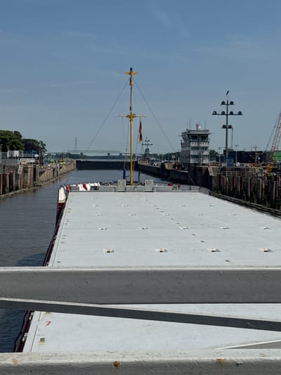 The image shows a view from the deck of a vessel, looking forward into a canal lock. The vessel is a barge or a similar cargo ship, with a large, flat, white deck covered in numerous square hatches. The canal walls are visible on either side, constructed of weathered wood pilings and stone. In the distance, a control tower with multiple windows and a control room on top stands by the lock. There are also navigational lights, a tall mast with a flag, and a crane visible further down the waterway. Trees and industrial structures can be seen on the left bank, and a large bridge crosses the canal in the far distance. The sky is clear and blue, indicating a sunny day. This scene is likely within the Brunsbüttel Elbe-Port, specifically the North Lock of the Kiel Canal, given the presence of the control tower and the canal infrastructure characteristic of this port. No people are visible in the image.