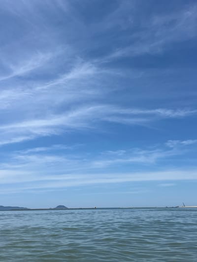 The image depicts a serene seascape with a vast expanse of blue sky filled with wispy white clouds. The horizon line is marked by a distant coastline with hills and a breakwater structure. The water in the foreground exhibits gentle ripples, reflecting the colors of the sky. Several small boats and what appear to be construction elements are visible along the distant breakwater. The scene suggests a tranquil day at the coast, likely in the vicinity of Hoi An, Vietnam, given the characteristic landscape and potential for such coastal features. There are no people or specific actions captured in the frame.