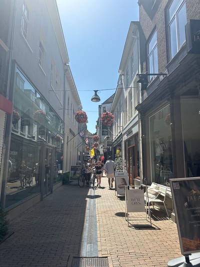 This is a vertical, eye-level shot of a cobblestone street lined with shops on both sides. The sun is shining brightly, casting long shadows, indicating it is likely midday. The scene is bustling with activity. Several people are walking along the street, some with bicycles. Hanging flower baskets add color and a cheerful atmosphere to the scene.

The shops on the left side of the street have large glass windows, reflecting the opposite buildings and the sky. One shop sign displays "MOONY" and "WE'RE OPEN". Another sign is partially visible with text "CLUSE". On the right side, a shop sign reads "TEAMSPORT Lee". Further down the street, more signs are visible, including "JA" and "JABA". The architecture of the buildings appears to be traditional European style with brickwork and tiled roofs. The overall impression is a vibrant and lively shopping street on a sunny day.