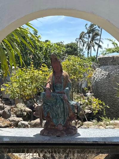 The image shows a statue of a seated figure, possibly a deity or an important historical person, positioned in a garden setting. The statue is made of a reddish-brown material with accents of teal and gold. It is seated on a rocky base. Behind the statue, there is lush green foliage, including palm trees and other plants. In the background, there is a large, textured, stone-like structure. The scene is framed by a white archway, suggesting it might be a decorative element within a larger structure or garden. The lighting suggests it is daytime, with a blue sky visible above the trees. The overall environment appears to be tropical or subtropical. The provided context mentions Tuy An Nam, Vietnam, which might indicate the geographical location of this scene.
