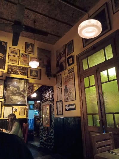 The image shows the interior of a bar or restaurant located in Victoria, Argentina, at the coordinates -34.452, -58.54. The scene is dimly lit, with a few ceiling lights casting soft shadows. There is a woman sitting in a booth near a window. The wall behind her is adorned with framed pictures and posters, including several depicting scenes of daily life in Argentina. There is a wooden bar in the background, with a doorway behind it. The room's ambiance is cozy and intimate, with the low lighting creating a warm atmosphere. The overall impression is one of a local establishment, frequented by residents and tourists alike.  The window in the back is covered by glass panes divided by wooden frames. There is an empty chair in the foreground, near a table with a napkin. The picture on the right side is a poster with a picture of a person, likely an artist,  in the center. The text on the poster is "Victoria,"  in a stylized font with the letters "VI" connected. The poster has a black background with white text and a picture.  The environment is warm and inviting.
