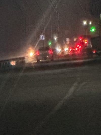 A blurry, slightly rotated aerial view shows city lights at night. Red, green, and white lights are strung out in a linear fashion, likely representing traffic or buildings. A road or structure appears as a dark, broad vertical band on the right side. The overall image is dark and somewhat grainy. A sign with the number "13" is visible.