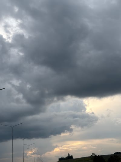 The image shows a street scene in Кояново, Russia, featuring a row of streetlights stretching into the distance. The sky is filled with dark, heavy storm clouds, suggesting an impending downpour. The lighting is dim, with a sliver of brighter sky visible near the horizon, possibly indicating the sun attempting to break through. The foreground is dominated by the stark silhouettes of the streetlights, emphasizing their repetitive nature along the road. A grassy embankment with a few scattered trees can be seen in the far distance, adding a natural element to the otherwise urban infrastructure. There are no people visible in the image. The overall atmosphere is one of a cloudy, possibly humid, day, characteristic of a storm's approach.