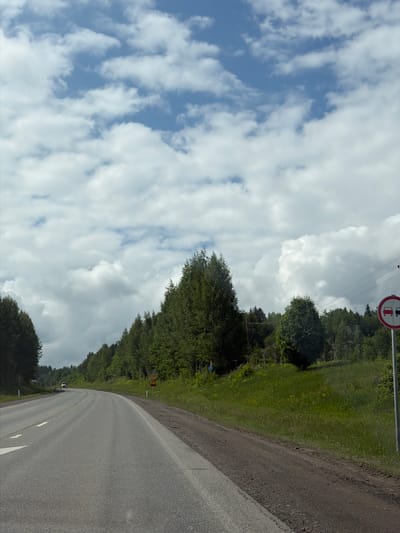 The image shows a rural road scene under a bright, partly cloudy sky. The asphalt road, marked with white dashed and solid lines, curves gently towards the left, receding into the distance. To the right of the road, there is a narrow dirt or gravel shoulder, followed by a strip of green grass and then a dense line of deciduous trees and foliage.
In the upper right corner, a circular traffic sign with a red border is clearly visible. It depicts two vehicles (a car and a truck) side-by-side, crossed out with a red diagonal line, indicating a "No Overtaking" zone for all motor vehicles. Above the trees and near this sign, power lines are also discernible. Further down the road, a smaller yellow diamond-shaped warning sign can be seen on the right, but its specific details are unclear due to the distance.
The sky is a mix of clear blue and scattered white cumulus clouds, suggesting fair weather during daylight hours. No people or specific activities are present, portraying a calm roadside environment in Башкултаево, Russia.