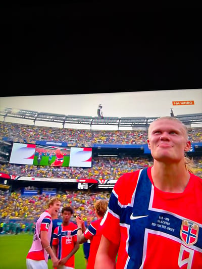 The image captures a scene from a soccer match, likely on a television screen due to the screen glare and the broadcast overlay. In the foreground, a male soccer player with blonde hair and fair skin is prominently featured, looking upwards and to his right with a slight smile. He is wearing a red and blue soccer jersey with the Norwegian flag motif. The jersey displays text and a crest: "FRA - NOR", "01.07.2026", and "NEW YORK NEW JERSEY", along with a crest that reads "NORGE". To the left of him, two other male soccer players in similar red and blue jerseys are visible. One is wearing a pink bib over his jersey and appears to be mid-sentence or shouting, while the other is looking towards the first player.
In the background, a large stadium filled with spectators wearing yellow and blue attire is visible. The stands are packed, indicating a significant event. On the field, some players are seen in the distance. There are large screens displaying images, one of which shows a player in a red uniform. A banner with orange text "НА ЖИВО" (meaning "LIVE" in Bulgarian) is present in the upper right corner of the screen.
The location described as the city of Ognianovo, Bulgaria, is not directly discernible from the visual elements of the image itself, as the context provided by the soccer match and stadium does not inherently point to a specific city in Bulgaria.
