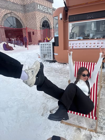 This image depicts an outdoor, snow-covered scene in Yeghipatrush, Armenia. In the foreground, the lower legs and feet of two individuals are visible. On the left, a person's left leg and foot, wearing black pants, a white sock, and a light-colored sneaker with a thick sole, are elevated and positioned to intersect the view of the second person's leg. On the right, a woman's legs, clad in black pants and a black chunky boot with a ridged sole, are extended. She is seated in a red and white striped deck chair. A dark-colored smartphone rests on a light-colored circular surface in the lower center.
In the midground, the same woman is visible, seated in the striped deck chair. She has long, dark hair styled in two braids, wears dark sunglasses, a white long-sleeved turtleneck top, and black pants. She is smiling and has both arms raised above her head, with her hands forming a "rock on" gesture. To her right, a brown, mobile food truck or kiosk is parked on the snow. It features a service window displaying a menu with various beverage options and a small security camera mounted near the top. A large, silver-colored ventilation grille is visible on its side.
In the background, a multi-story brick building with intricate architectural details, including arched windows, stands prominently. The word "ANGELINE" is displayed in large, white letters on its facade. A red mesh fence is visible in front of the building, behind which several other individuals are seated on bean bags or similar low seating, alongside what appears to be ski equipment. The entire ground surface is covered in snow, under an overcast sky.