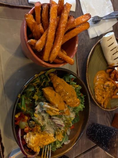 A close-up, overhead shot displays a meal on a wooden table. To the left, a bowl of sweet potato fries is partially visible, with some fries spilling out. In the center, a dark bowl holds a salad topped with chicken and a creamy dressing, with red onion and olives scattered throughout. To the right, a plate features a wrap and more chicken with fries. A fork rests on a white napkin near the fries. The meal appears to be set at a restaurant in Peterborough, United Kingdom, although no specific establishment is identified. The lighting suggests an indoor setting.