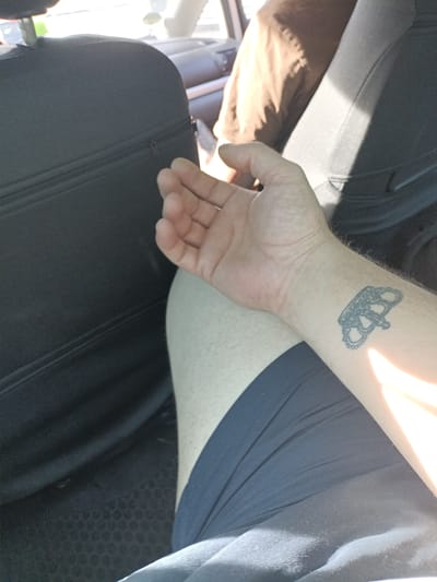 The image shows a person's arm with a tattoo of a crown on their wrist. The person is sitting in what appears to be the back seat of a car. The interior of the car is visible, including a black car seat and a glimpse of another seat with a person wearing a brown jacket. The sunlight streaming into the car suggests it is daytime, possibly a sunny day. The setting appears to be casual, with the person wearing dark shorts. There are no visible signs or text in the image. The image appears to have been taken in Vranov, Czechia.