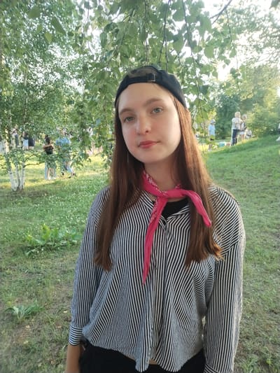 A young Caucasian female is standing outdoors in a park-like setting, looking towards the camera. She is wearing a black baseball cap, a black and white striped long-sleeved shirt, and a bright pink bandana tied around her neck. Her long brown hair falls down her back. In the background, several other people are visible, some appearing to be children, and trees and grass surround the scene. The lighting suggests it is daytime, possibly late afternoon, given the warm glow. The location is Detskii Dom (Bashenino), Russia.