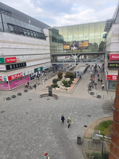 An elevated, wide-angle daytime photograph depicts an urban commercial and pedestrian area in Nyíregyháza, Hungary, specifically the plaza adjacent to the Korzó Shopping Center and the Móricz Zsigmond Theater, with Dózsa György út visible beneath a skywalk. The sky is overcast.
The background features a multi-story modern building with light-colored stone cladding and extensive dark-tinted glass windows. Its ground level houses retail establishments including CSL Plasma, MOHITO, SPAR, DAK, and DEICHMANN. A prominent glass pedestrian skywalk connects two building sections over Dózsa György út. Behind the skywalk, a large digital billboard on a glass facade displays theatrical advertisements in Hungarian.
The foreground and mid-ground consist of a gray paver-tiled pedestrian plaza. This area incorporates several square planters with small trees and shrubs, along with a larger central landscaped island containing similar plantings and white gravel. A curved public bench with brown wooden seating and an integrated information kiosk is situated on the right side of the plaza. Multiple bicycles are parked along the pedestrian pathways, some secured to bollards. Manholes and drainage grates are visible in the paving. A narrow strip of ground with bare trees and shrubs runs along the right edge, adjacent to a partial brick wall. A grey trash receptacle and a white directional signpost, indicating "NYÍREGYHÁZA" among other locations, are positioned on the right.
Approximately 20-30 individuals, dressed in casual attire suitable for cool weather, are observed walking across the plaza and along the pathways. Two pedestrians are in the immediate foreground walking away from the viewer, one in a dark jacket carrying a white bag, and another in a light green jacket carrying a white object. A third individual in a red hat and light jacket is visible in the bottom center. Pedestrian and light vehicular traffic, including a white concrete ramp, are visible on Dózsa György út beneath the glass skywalk. Storefronts for Hervis and BEST are visible on the right side of the street level.