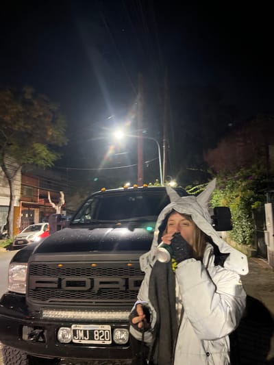 The image depicts a woman in a gray hooded jacket holding a can, standing next to a black Ford truck with license plate "JMJ 820". The location appears to be a street at night, with streetlights visible. Buildings and trees are also visible in the background. The scene is possibly in Olivos, Argentina, based on the license plate mentioning "Argentina".