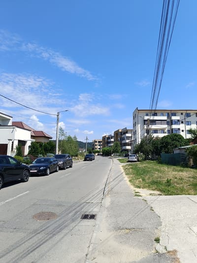 The image shows a street scene in Târgu Jiu, Romania, on a sunny day with a blue sky and scattered clouds. The foreground features a paved road with tire tracks and two manhole covers. Several cars are parked along the side of the road, including a black SUV, a black Audi A4 sedan, and a grey SUV. Residential buildings and apartment blocks line the street, with modern architecture evident in the apartment buildings. Trees and greenery are present along the roadside and between the buildings. Overhead, power lines are visible against the sky. The overall impression is a typical urban residential street.