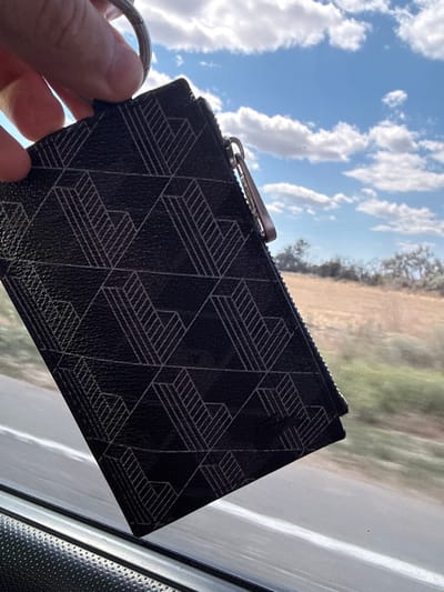 A person's hand holds a black zippered cardholder with a geometric pattern of white lines. The cardholder is attached to a silver keyring. In the background, a blue sky with white clouds is visible, along with a dry, grassy field and trees. The image appears to be taken from inside a vehicle, as indicated by the presence of a window reflecting the sky and landscape, and a black textured surface that could be part of a car's interior. The specific location of Vyzyrka, Ukraine, is not determinable from the visual information in the image.