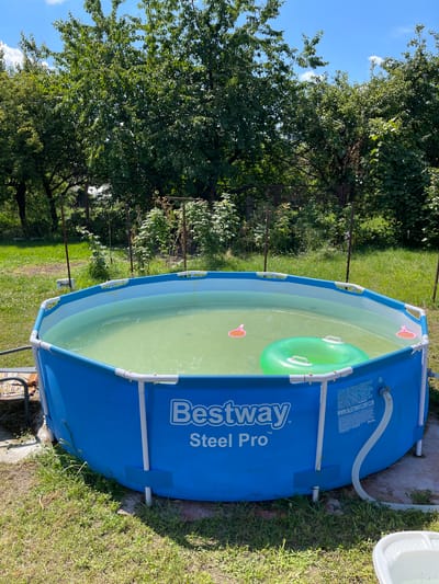 In the village of Ivankovychi, Ukraine, a bright blue, above-ground Bestway Steel Pro pool is filled with murky green water under a clear blue sky with scattered clouds. A vibrant green inflatable ring floats near the center of the pool, accompanied by two small, pink toy devices. The pool is situated on a patch of overgrown green grass, bordered by more lush vegetation and mature trees. A white hose snakes away from the pool, and a white plastic tub sits to the right. The scene suggests a warm, sunny day, likely in a residential backyard.