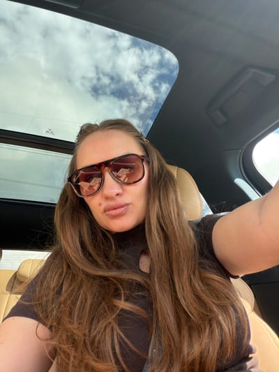 The image shows a woman taking a selfie in a car. She has long, brown hair and is wearing sunglasses with tortoise-shell frames and a dark brown top. The car has a sunroof, through which a cloudy sky is visible. The interior of the car appears to be made of tan leather. The photo is taken from a slightly high angle, looking down at the woman. There is no visible indication of the location in Severny, Russia.