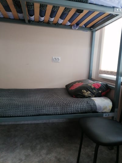 The image shows a close-up, eye-level view of a bunk bed in a room, likely a dormitory or hostel, in Komsomolsky, Russia. The lower bunk is made up with a grey, textured blanket and two pillows. One pillow has a dark floral pattern with red roses. A small, black stool with three legs is positioned in front of the lower bunk. The bunk bed frame is made of grey metal, and the upper bunk's wooden slats and bedding are visible. A light-colored wall with a textured surface is behind the bed. An electrical outlet with two sockets is mounted on the wall to the left of the bed. A window with sheer blinds is visible to the right, suggesting daytime. The overall impression is of a simple, functional living space. There is no visible text in the image.