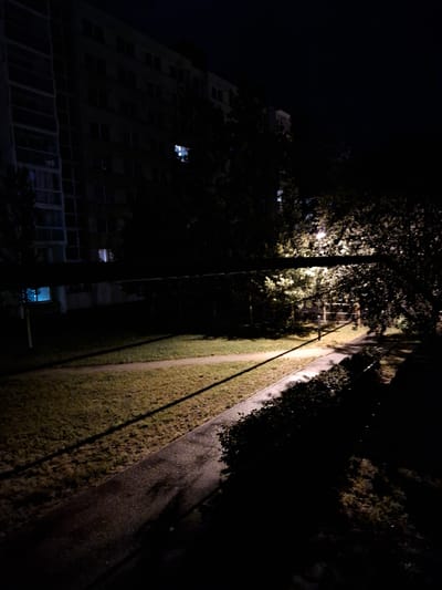The image depicts a nighttime scene outside a residential building in České Budějovice, Czechia. A tall, multi-story apartment building is visible on the left, with several windows illuminated. The building appears to be constructed from concrete and glass. In front of the building, a path or sidewalk curves through a grassy area. A street lamp illuminates a portion of the path and surrounding foliage, casting long shadows. Trees with dense foliage are present, some of which overhang the path. A horizontal pipe or conduit runs across the middle of the frame, obscuring a portion of the view. The overall impression is one of a quiet, urban environment at night.