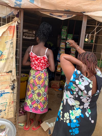 In Mape, Nigeria, two women are seen at a small market stall. The woman on the left, facing away from the camera, wears a red and pink heart-patterned tank top and a long, colorful African print skirt with circular motifs. She stands on red flip-flops. In front of her, a yellow basin and a box of onions are visible. To the right, another woman, also facing away, is dressed in a black dress with a floral pattern of blue, green, and white flowers. Her hair is styled in dark braids with red highlights, and she is reaching up to adjust something with her right hand, while her left arm is bent behind her head. The market stall has shelves stocked with packaged goods, and a metal gate with a padlock is part of the structure. A torn, yellow and white tarp hangs over the top of the stall. The ground appears to be dirt or concrete.