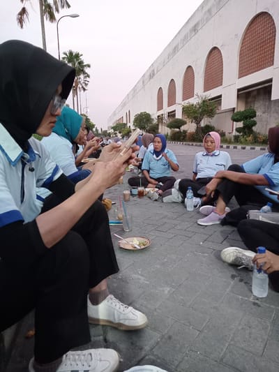 A group of approximately seven women are seated on a paved surface, possibly a parking lot or walkway, in what appears to be an outdoor setting. The women are wearing light blue shirts with dark blue collars and long black pants. Many are wearing hijabs of various colors. Several women are using cell phones, while others are eating or drinking. Empty plates, cups, and water bottles are scattered around them on the pavement. In the background, a large white building with arched windows and several trees and shrubbery are visible. Palm trees and street lights are also present in the background, under a dusk or overcast sky. Based on the evidence that the location is Sukomoro, Indonesia, the scene depicts a casual social event or a meal break outdoors for these women.