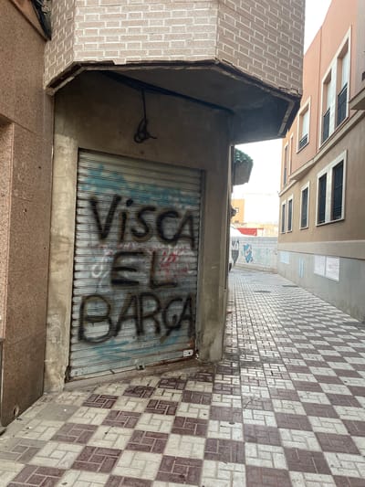 The image depicts a narrow street in La Línea De La Concepción, Spain, with a prominent, closed metal shutter on the left, covered in graffiti. The graffiti appears to say "Visca El Barca". The shutter is part of a building with textured, reddish-brown facade panels to its left and a concrete pillar. Above the shutter, a damaged concrete overhang juts out, showing exposed interior materials and electrical conduits. The ground is paved with a checkerboard pattern of light and dark red square tiles. The street opens up to the right, revealing more buildings. The building on the right has a beige facade with multiple windows, each featuring black bars and white shutters or frames. The street continues into the distance, where more buildings are visible, and a white wall with blue graffiti, including a shield-like emblem, can be seen. The sky is overcast, and the lighting suggests daytime. There are no people visible in the image.