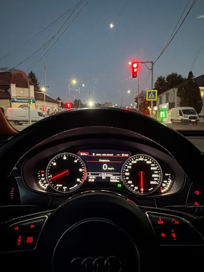 The image shows a view from the driver's seat of a car, looking at the dashboard and the road ahead. The car is stopped at a traffic light, which is red. The dashboard displays the car's speed as 0 km/h and the current temperature as 24.5°C. The car's odometer shows 163,608 km. In the background, there are buildings, streetlights, and other vehicles, including a white van. A pedestrian crossing sign is visible on the right side of the road. The sky is dark, indicating it is either dawn or dusk. The location appears to be in a city, likely Orhei, Moldova, given the presence of the "Victoriabank" sign which is a known bank in Moldova. The overall scene suggests a moment of pause in urban traffic during low-light conditions.