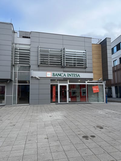 The image depicts the exterior of a modern commercial building located at Bulevar Nemanjića 13, Niš, Serbia. The primary subject is a Banca Intesa branch situated on the ground floor.
The two-story building features a facade predominantly clad in large, light grey rectangular panels. The upper floor exhibits several wide rectangular windows, each equipped with external horizontal aluminum louvers or shades. A section on the upper right of the building is clad in light brown, horizontally oriented, wood-look panels.
At ground level, the central section houses the Banca Intesa entrance, consisting of two clear glass doors. Above the doors, the "BANCA INTESA" logo is displayed in green text with an orange graphical element. A small, white security camera is mounted below the logo, to the left. Through the large glass window to the right of the entrance, the interior of the bank is visible, characterized by red walls or accents and two automated teller machines (ATMs). A red, square-shaped sign with white text "ZONA 24h" (24-hour zone) is affixed to the interior surface of this window.
To the immediate left of the Banca Intesa entrance, another glass door provides access to an internal staircase ascending upwards. A "No Smoking" symbol sticker is present on this door. To the far right of the bank's main window, a blue fire hydrant is visible on the paved area.
The foreground consists of an expanse of light grey square concrete paving stones, showing some scattered dark stains and discoloration. No individuals, vehicles, or significant activity are present in the frame. The sky overhead is overcast.