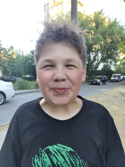 The image shows a close-up, outdoor portrait of a young boy with messy brown hair, squinting his eyes and puckering his lips. He is wearing a black t-shirt with a green design. The background suggests an urban setting, with trees, a building, and parked cars visible. The lighting indicates it is daytime, possibly late afternoon or early evening.